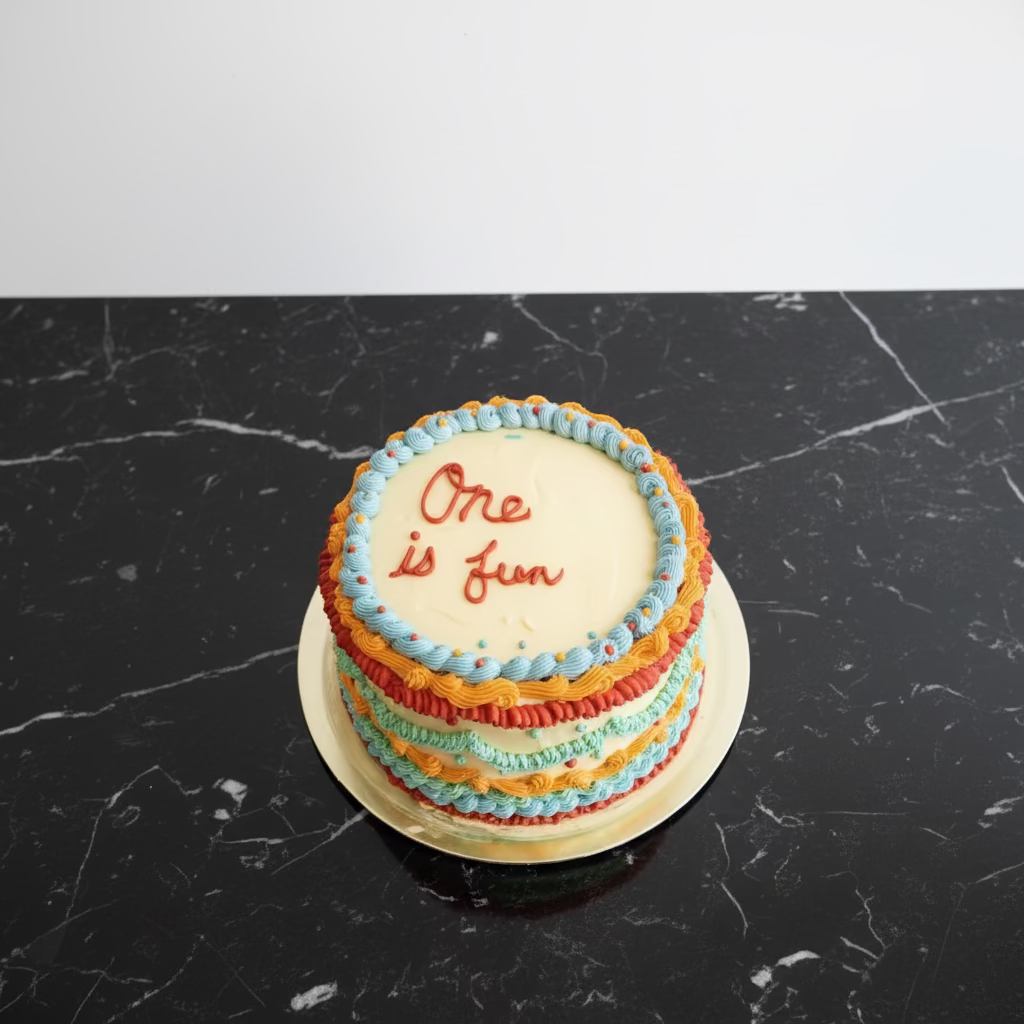 A multi-layered birthday cake with colorful piped frosting borders and the words 'One is fun' written on top, sitting on a kitchen counter surrounded by baking tools and ingredients.